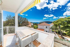 A balcony with a white table and wicker chairs, overlooking a coastal village with houses and trees. - Apartments Bozo (Okrug)