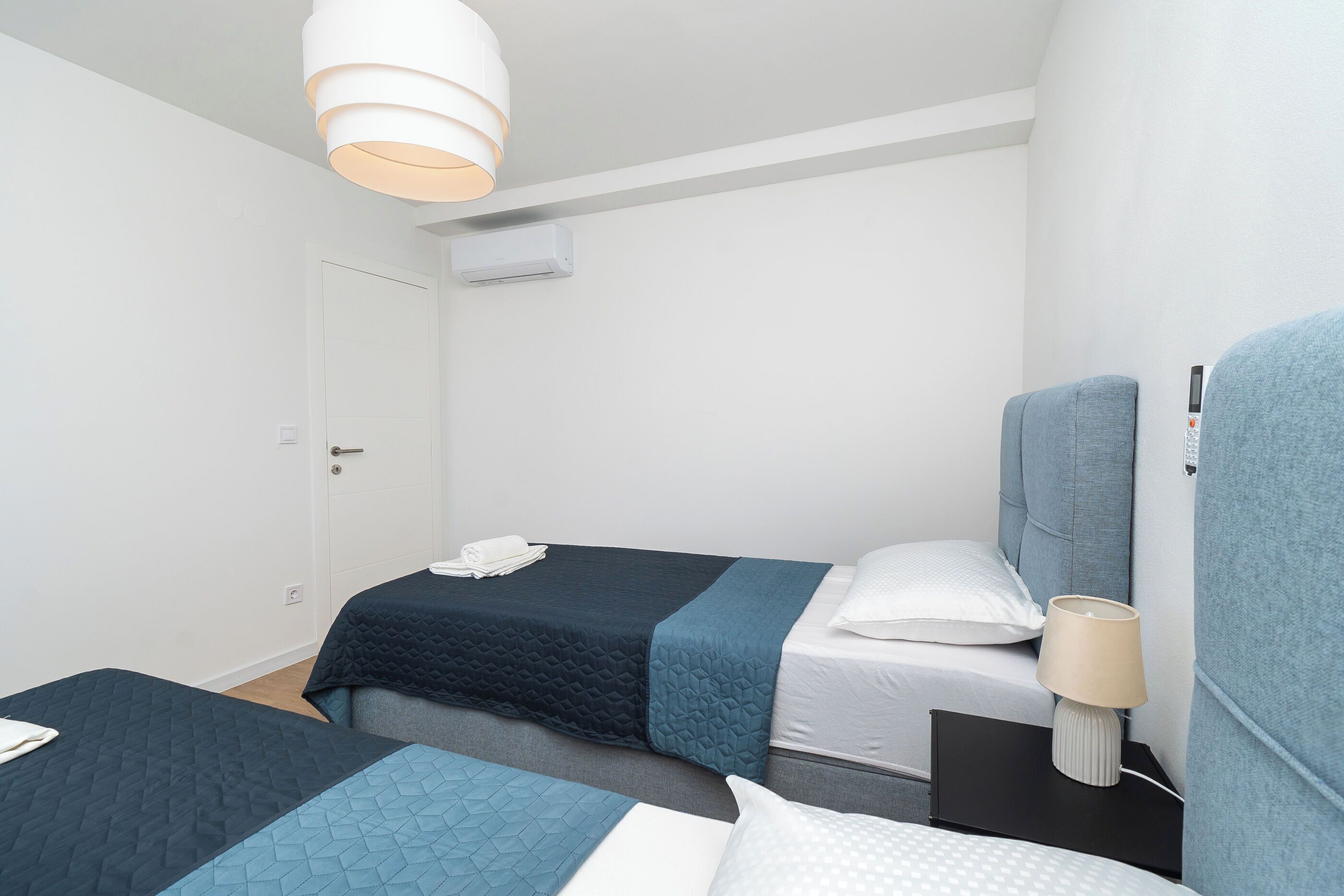 A hotel room with two beds, a bedside table with a lamp, a door, and an air conditioning unit on the wall.