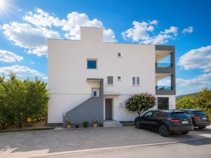 A modern two-story building with a flat roof, a small entrance, and a parking area with two cars. - Apartments Lenolu (Vodice)