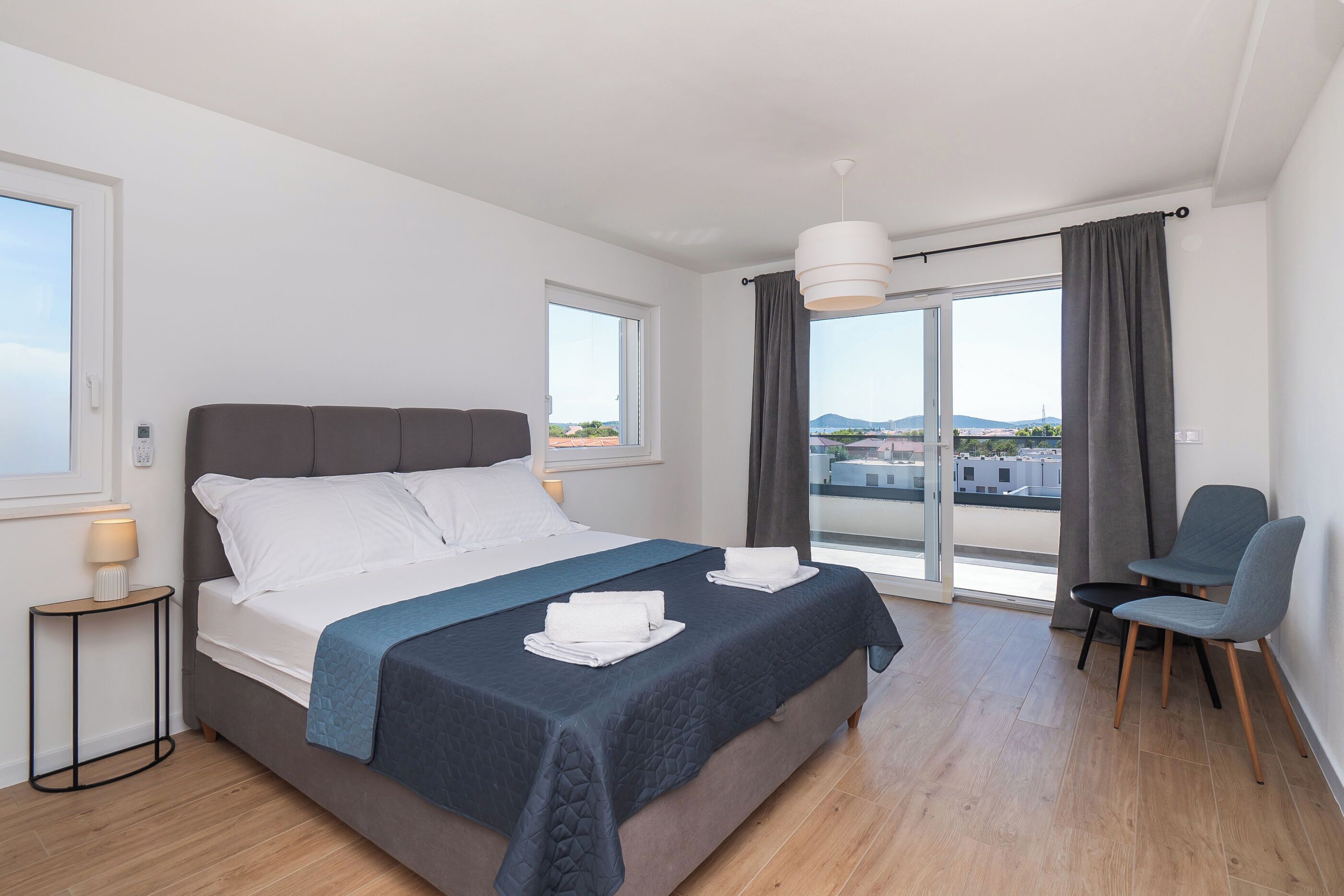 Quarto de hotel moderno com uma cama grande, uma mesa pequena com luminária, uma cadeira e vista para a cidade através de amplas janelas.