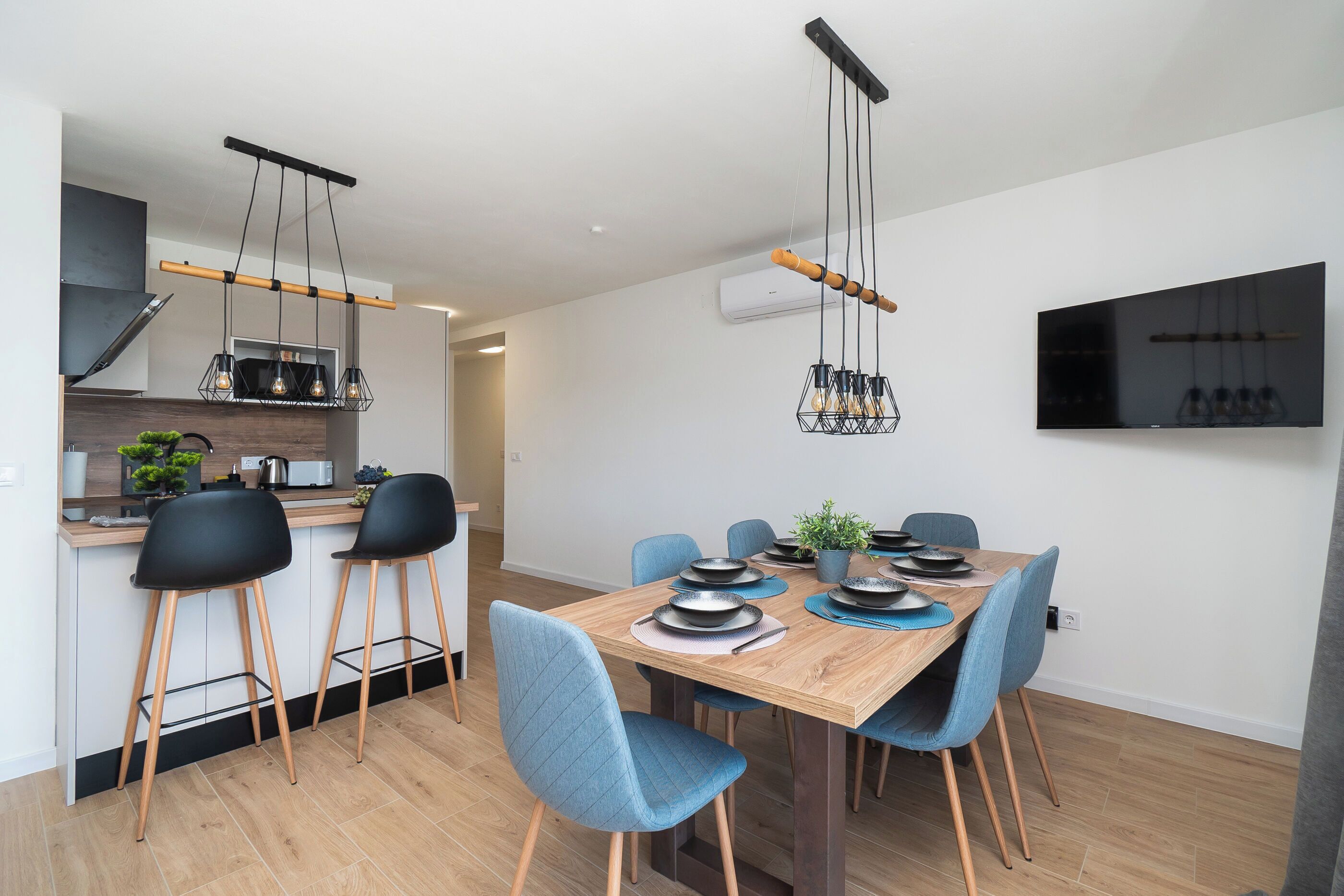 A modern kitchen with a wooden countertop, black bar stools, and a dining table set with plates and a potted plant.