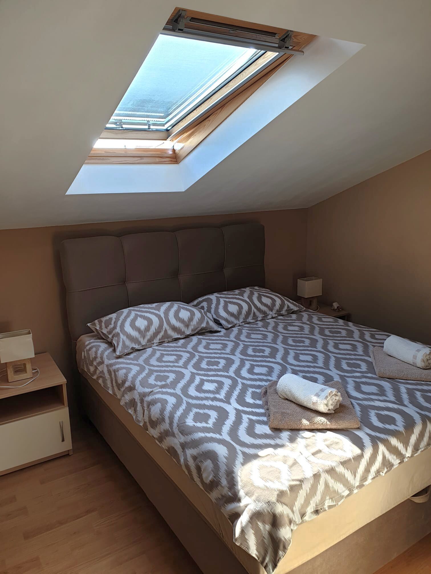 Une chambre avec un puits de lumière, un lit avec une literie à motifs, une table de chevet avec une lampe et une commode.
