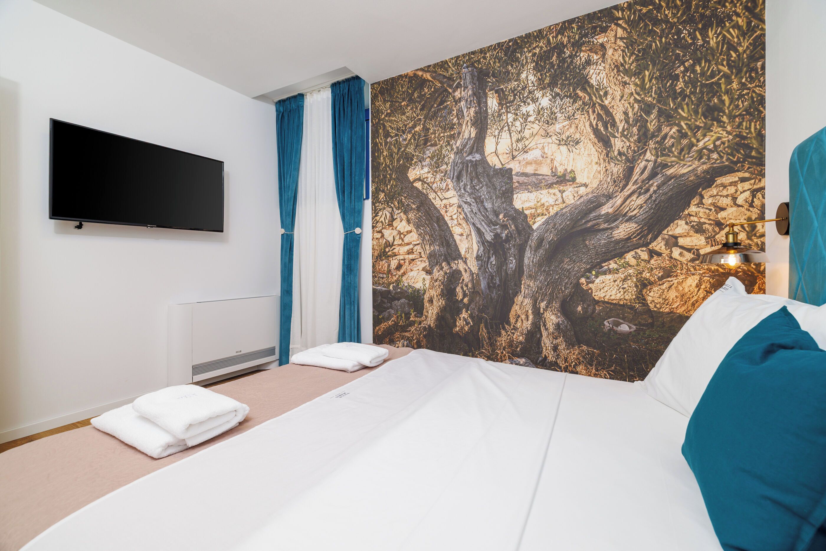 A neatly made bed with white linens, a flat-screen TV mounted on the wall, and a large tree mural on the wall behind the bed.