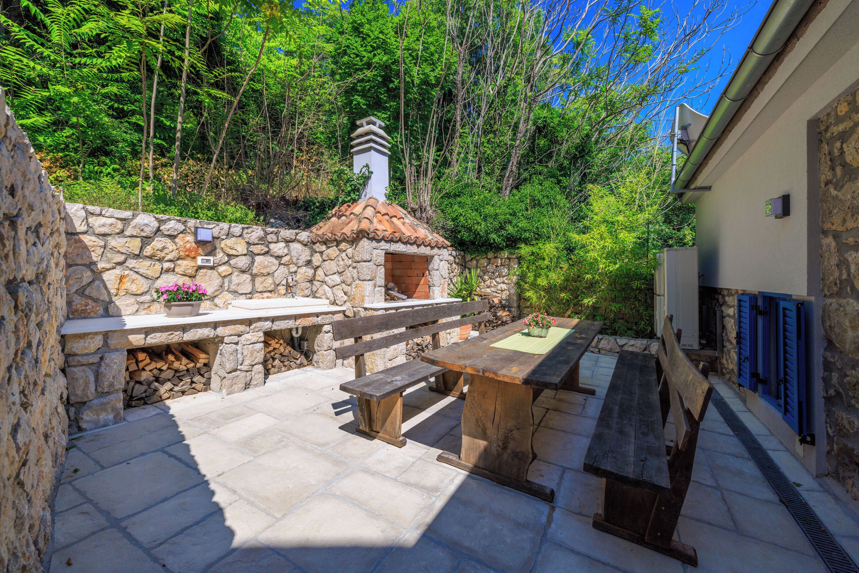 A stone patio with a wooden table and benches, surrounded by a stone wall and trees.