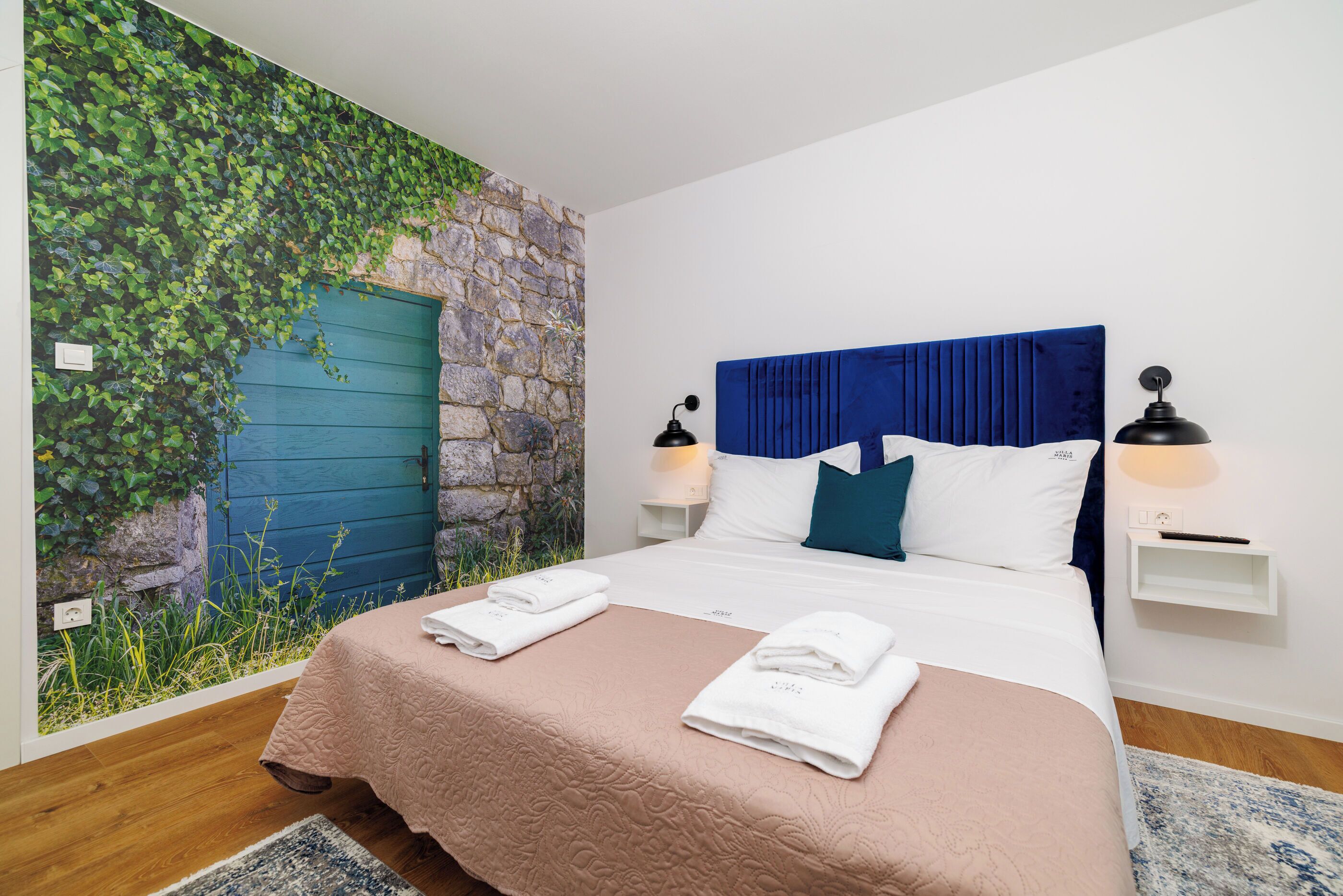 Een slaapkamer met een stenen muur, een blauwe deur, een bed met een blauw hoofdeinde, witte kussens en twee bedlampjes.