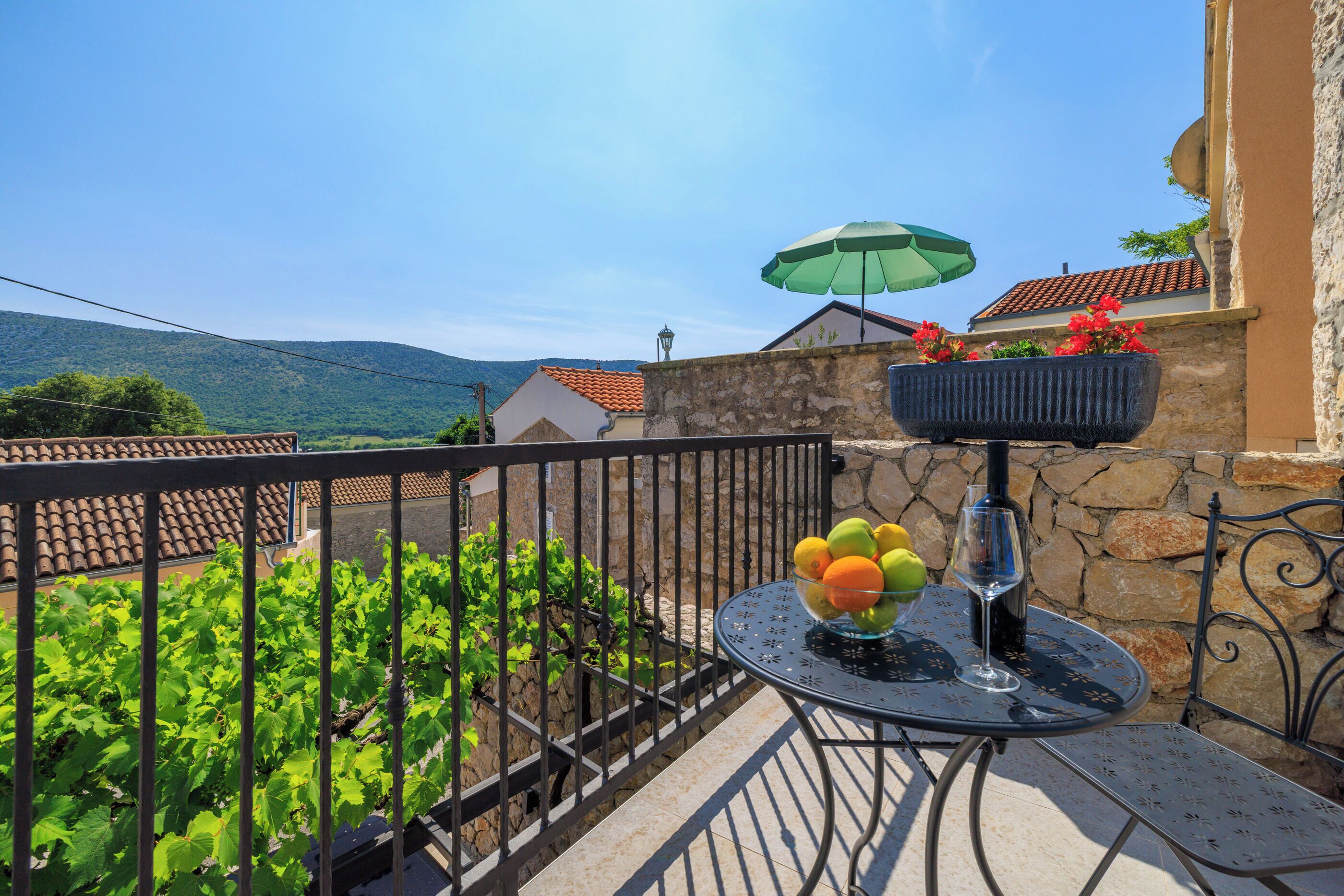A balcony with a table, a hanging planter with flowers, and a view of the countryside.
