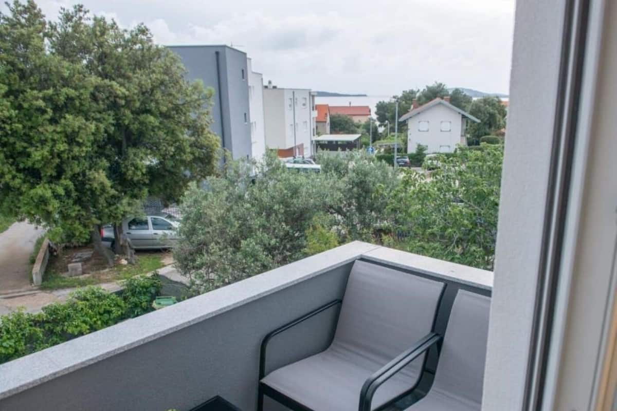 A balcony with a cushioned chair and a small table, overlooking a residential area with trees and buildings.