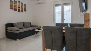 Ein modernes Wohnzimmer mit einer Couch, einem Esstisch mit Stühlen, einem kleinen Beistelltisch mit Pflanze und einem an der Wand montierten Flachbildfernseher.