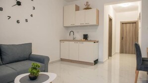 Ein modernes Wohnzimmer mit einer grauen Couch, einem kleinen runden Tisch mit einer Pflanze sowie einer Küchenzeile mit weißen Schränken und einer Kaffeemaschine.