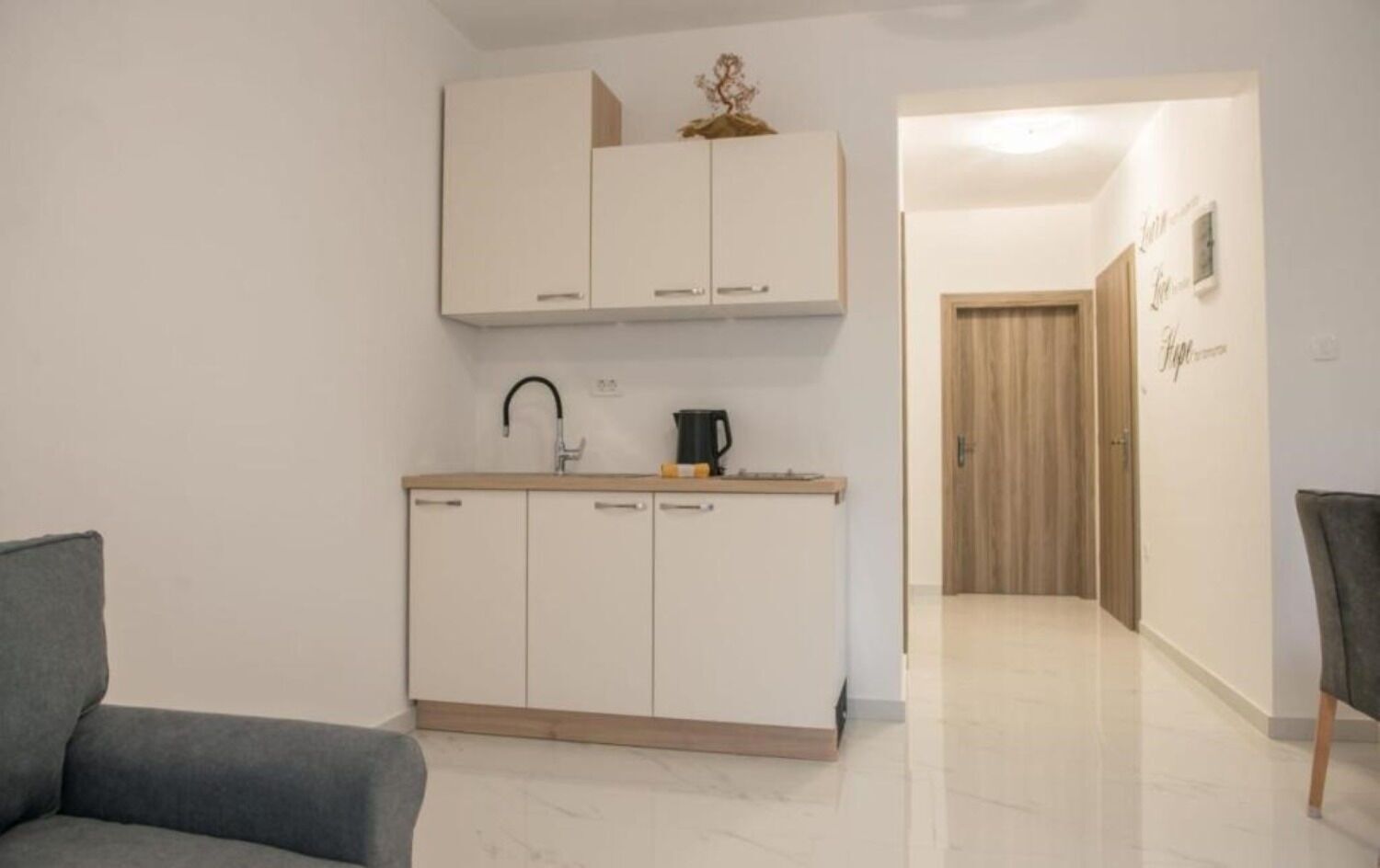 A compact kitchen with white cabinets, a sink, and a coffee maker. There is a grey sofa in the foreground and a wooden door in the background.