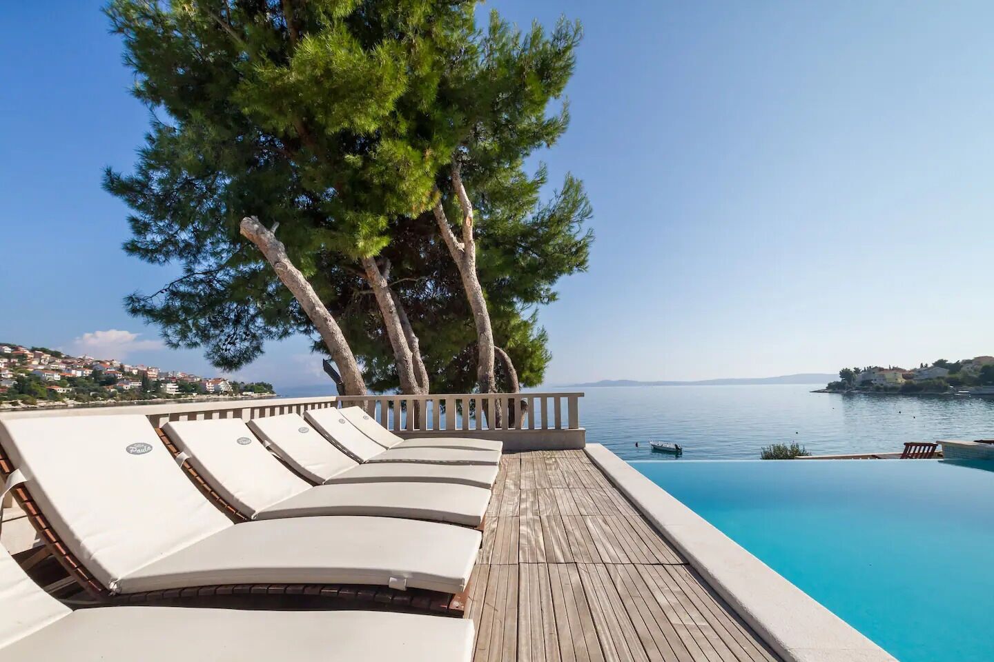 Eine Holzterrasse mit Liegestühlen mit Blick auf ein klares, blaues Schwimmbecken und eine malerische Aussicht auf das Meer und das entfernte Land.