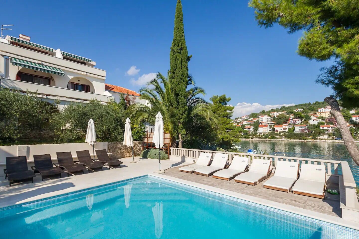 A swimming pool with lounge chairs and umbrellas, surrounded by greenery and a view of a lake and buildings.