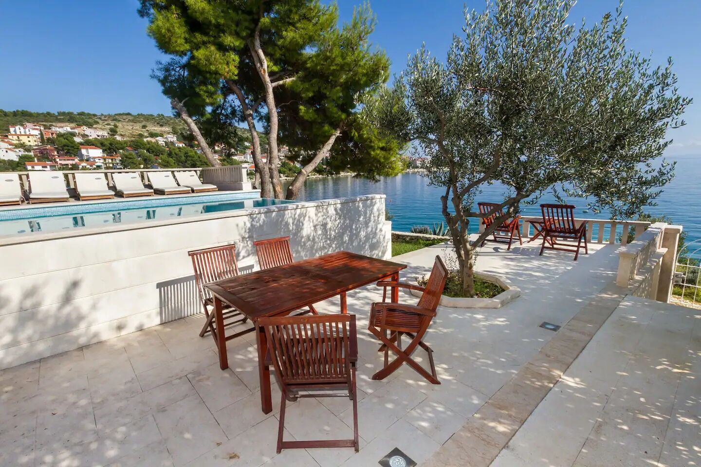 A poolside area with a wooden table and chairs, a small tree, and a view of the sea.