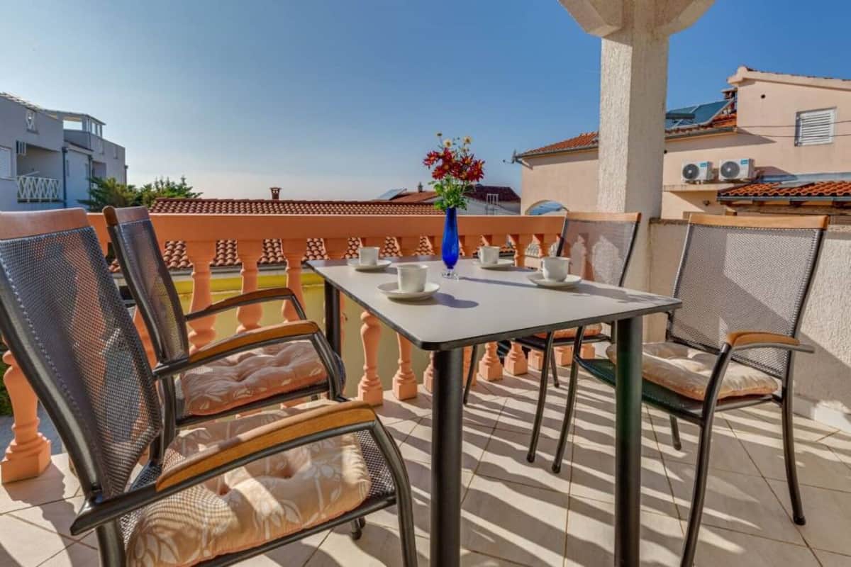 A terrace with a table and chairs, a vase with flowers, and a view of buildings in the background.