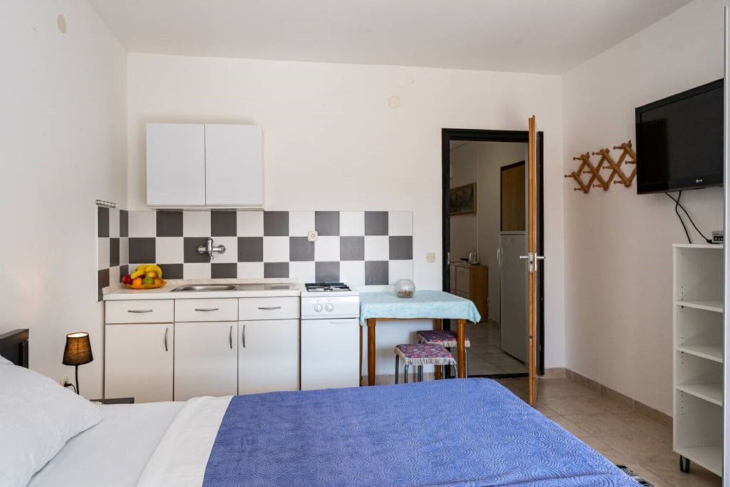 A bedroom with a bed, a small kitchenette, a table with a chair, and a TV mounted on the wall.
