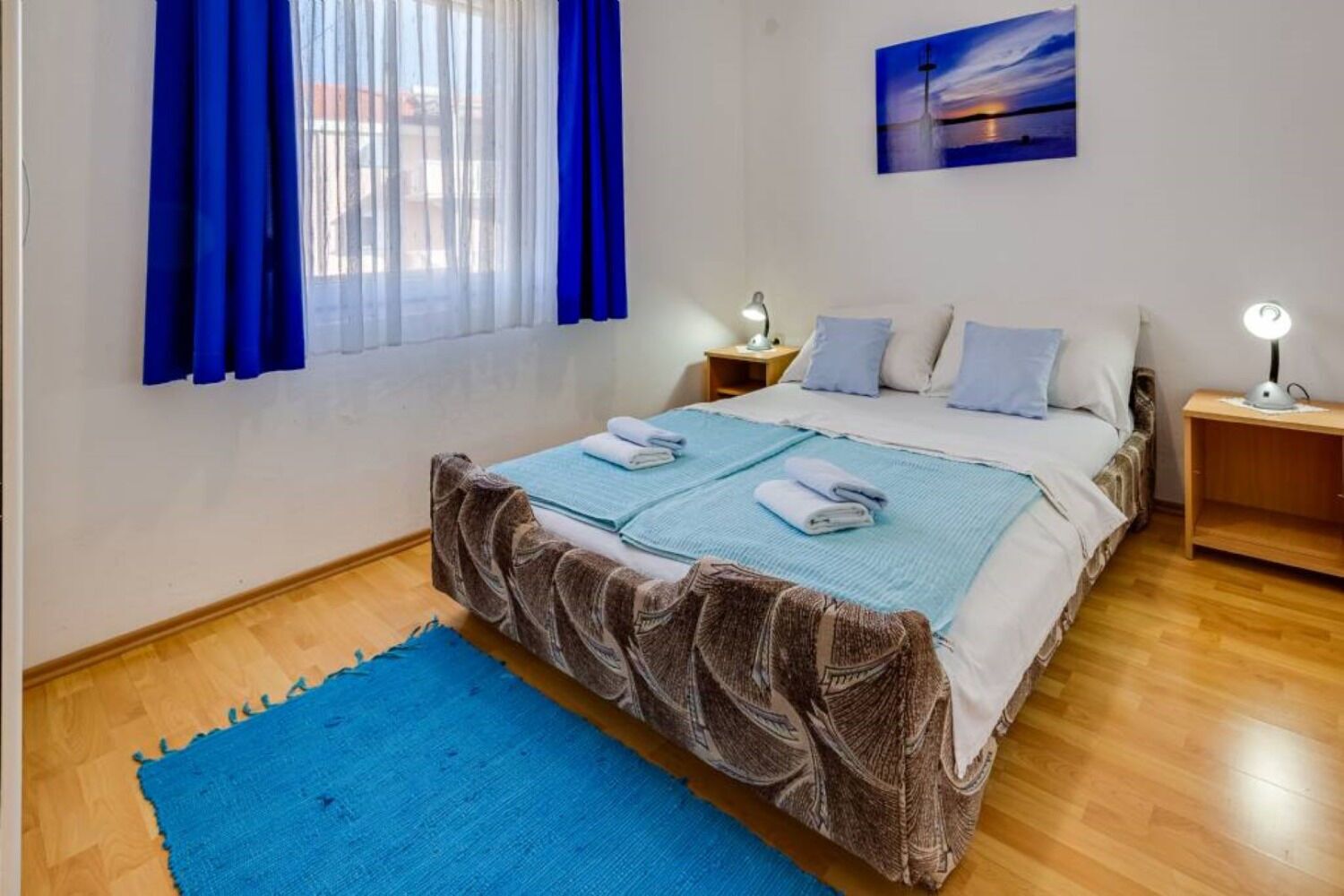 A bedroom with a bed, two bedside tables, a nightstand, a blue rug, and a painting on the wall.