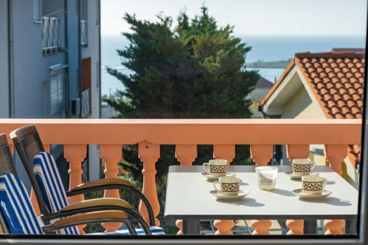 A balcony with a table set for tea, chairs, and a view of the sea and buildings.