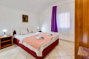 A bedroom with a bed, two bedside tables with lamps, a window with purple curtains, and a painting on the wall. - Apartments Tanja (Pakostane)