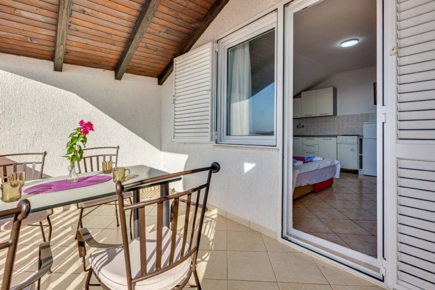 Ein kleiner, gut beleuchteter Raum mit Essbereich, der einen Tisch und Stühle bietet, einer durchgehenden Tür erkennbaren Küche und einem Balkon mit Holzboden.