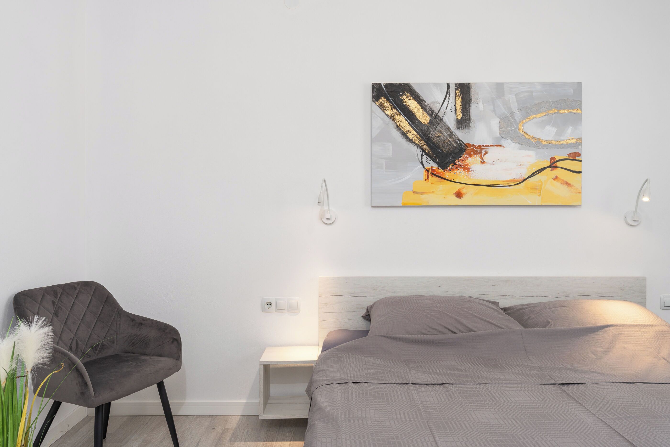 A modern bedroom with a bed, a nightstand, a chair, and a wall-mounted abstract painting.