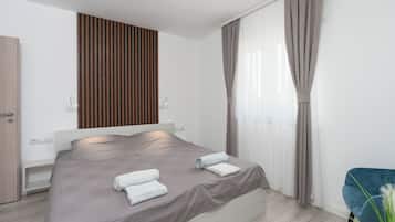 A modern hotel room with a large bed, a wooden panel wall, a window with curtains, and a blue chair.