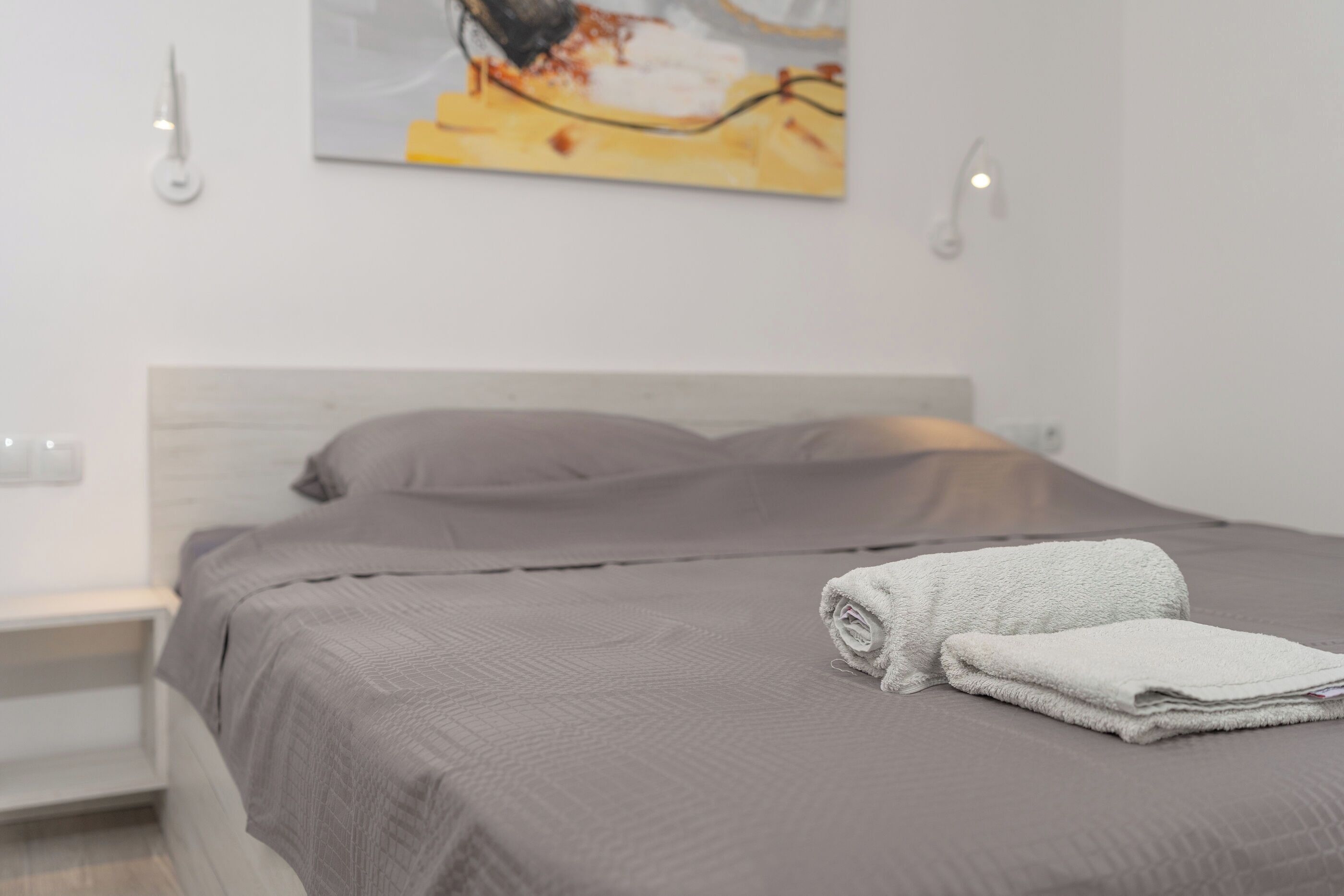 A neatly made bed with a folded towel and a pillow, a bedside table, and a wall-mounted light fixture.