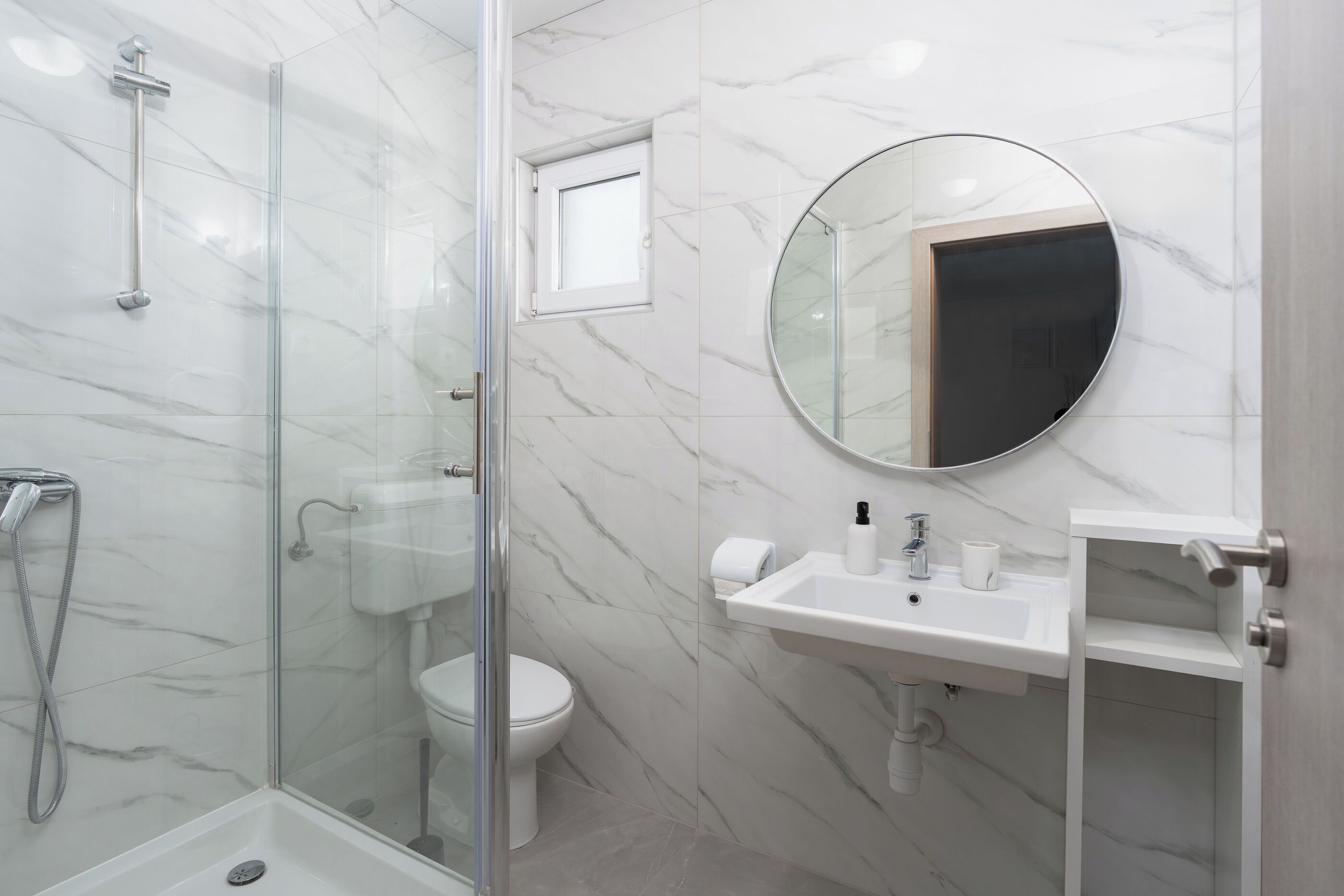 Un baño con ducha de mampara, inodoro, lavabo con espejo y una pequeña ventana.