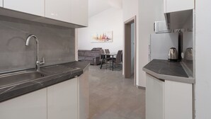 A modern kitchen with a built-in sink, a microwave, and a kettle on the countertop. There is a dining area with a gray sofa and a table in the background. - Apartment Juli (Vir)