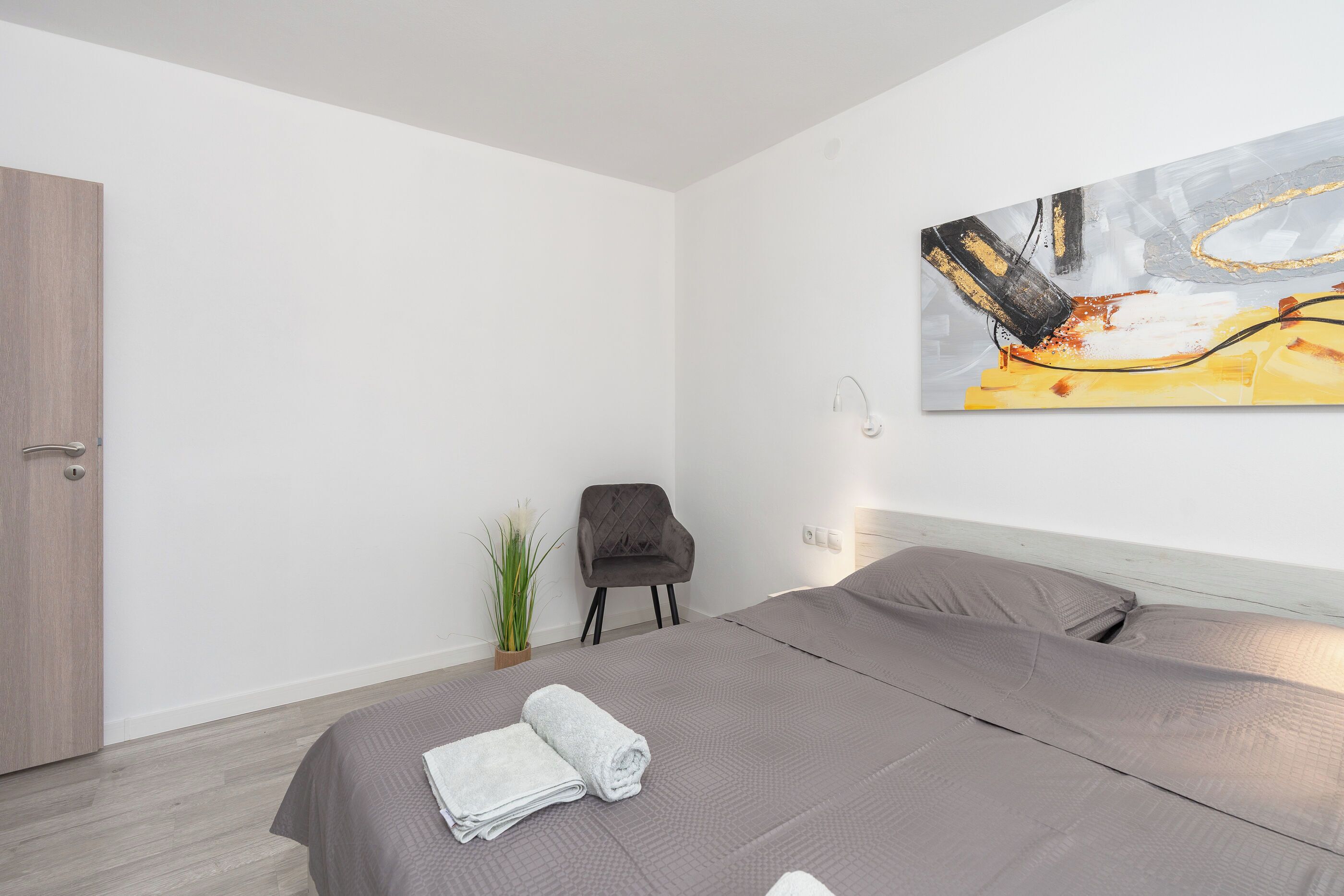 Habitación de hotel moderna con una cama grande, cabecera gris, un cuadro en la pared y una pequeña planta en maceta.
