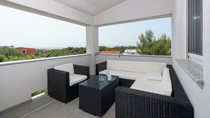A balcony with a sofa and two chairs, a glass-top table, and a view of greenery and buildings. - Apartment Juli (Vir)