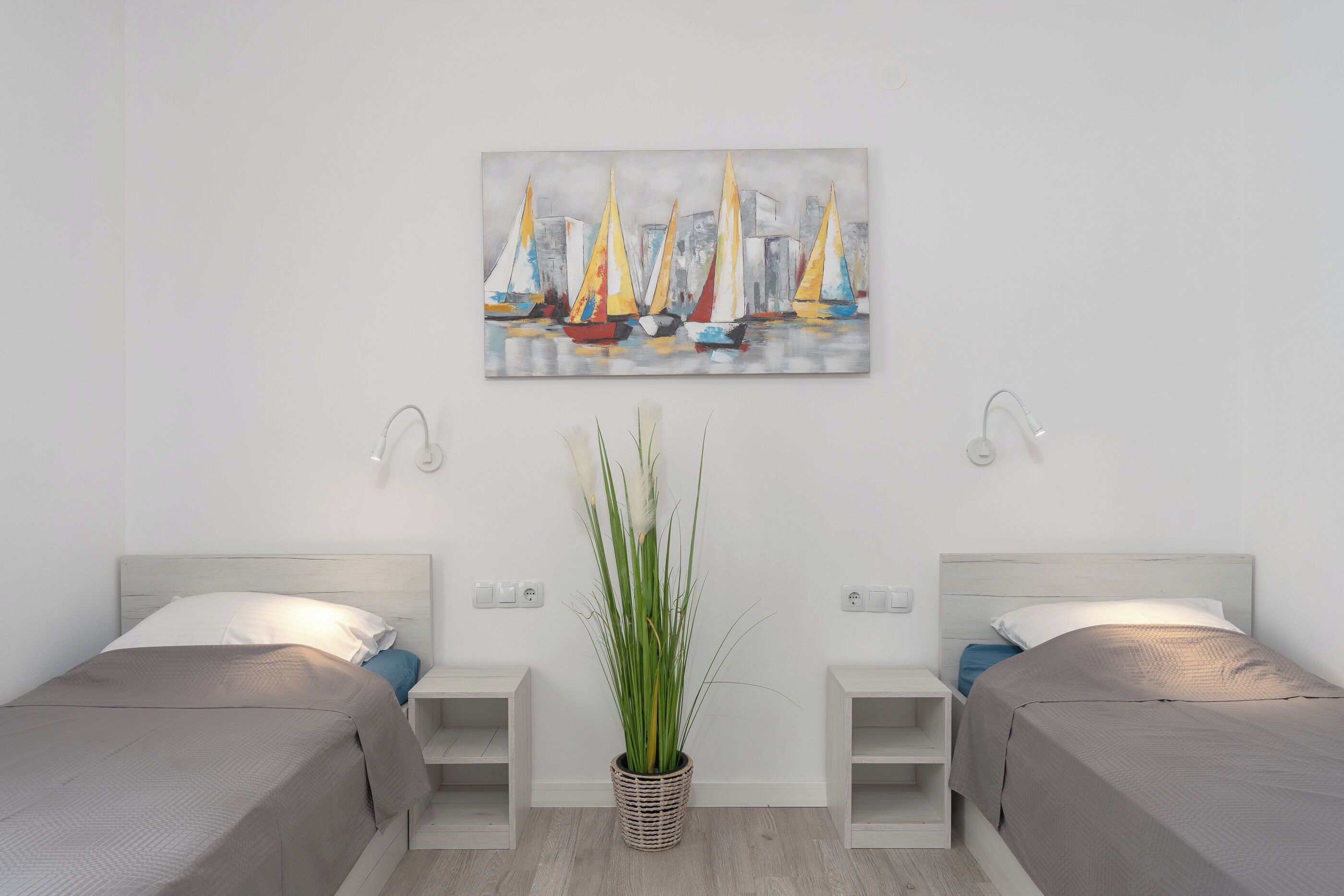 Habitación de hotel con dos camas, una mesita de noche con lámpara, una planta en maceta y un cuadro de veleros en la pared.