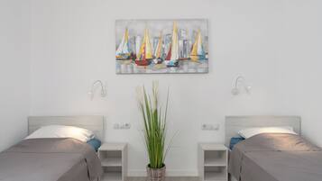 Habitación de hotel con dos camas, una mesita de noche con lámpara, una planta en maceta y un cuadro de veleros en la pared.