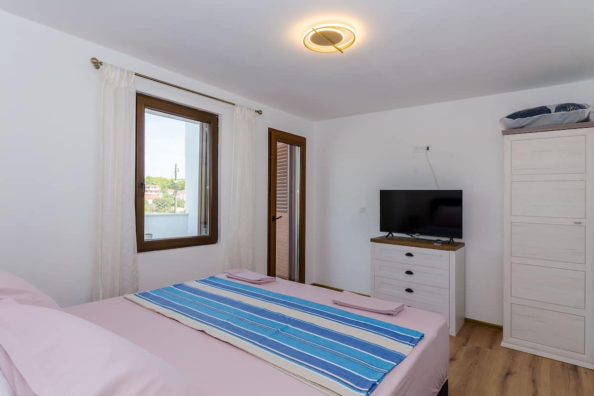 A bedroom with a bed, a television on a stand, a wardrobe, and a window with curtains.