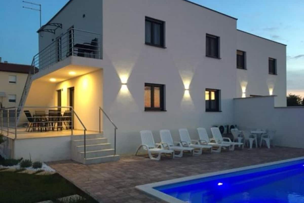 Un edificio moderno de dos pisos con piscina, mobiliario exterior y una escalera que lleva a un nivel superior.