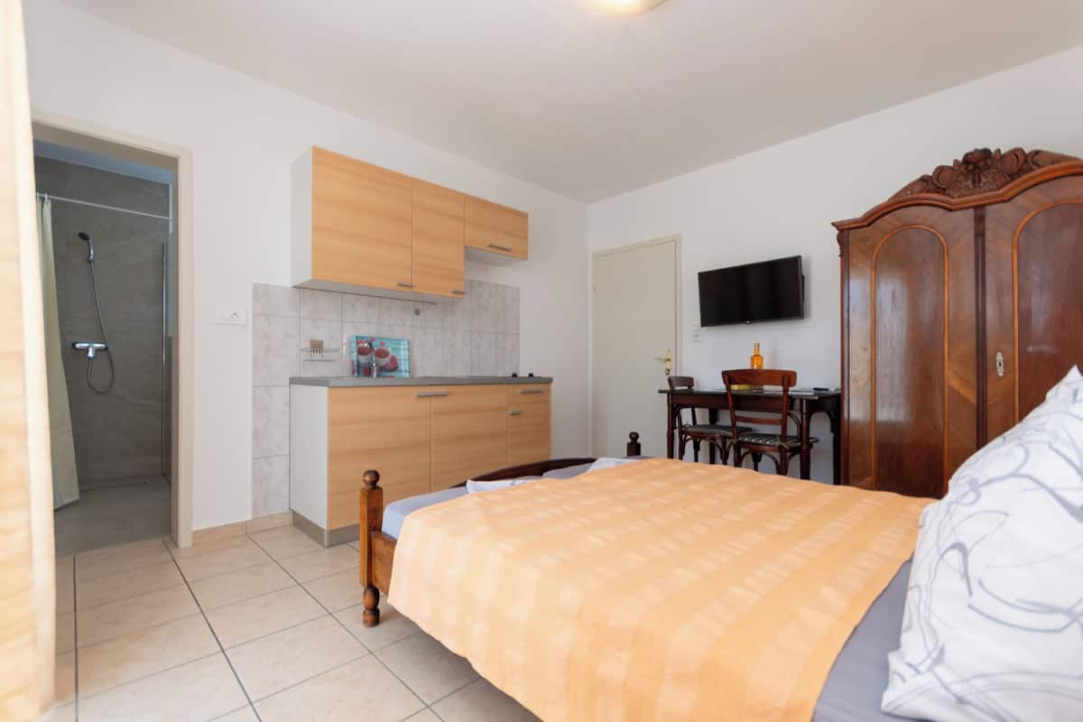 A bedroom with a bed, wooden wardrobe, a desk with a chair, a television mounted on the wall, and a bathroom with a shower.