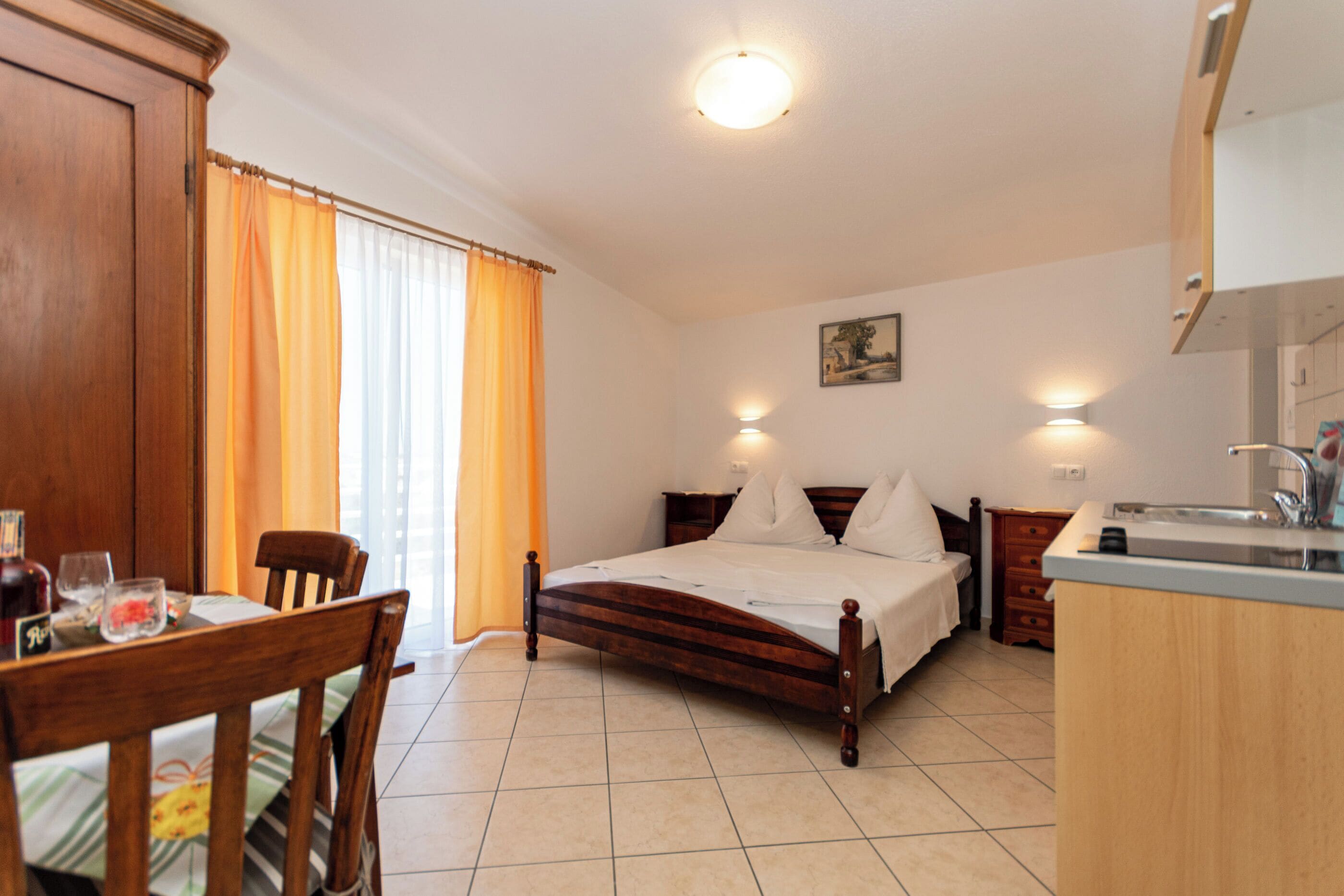 A bedroom with a wooden bed, a dining table with chairs, a kitchen area with a sink, and a window with curtains.