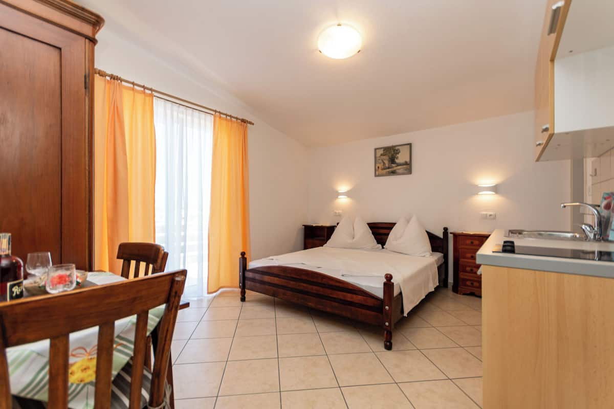 A bedroom with a wooden bed, a bedside table, a nightstand, a window with curtains, and a kitchen area with a sink and cabinets.