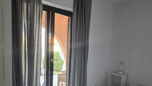 Een slaapkamer met een bed, nachtkastje, raam met gordijnen en uitzicht op een buitengebied met een stoel en tafel.