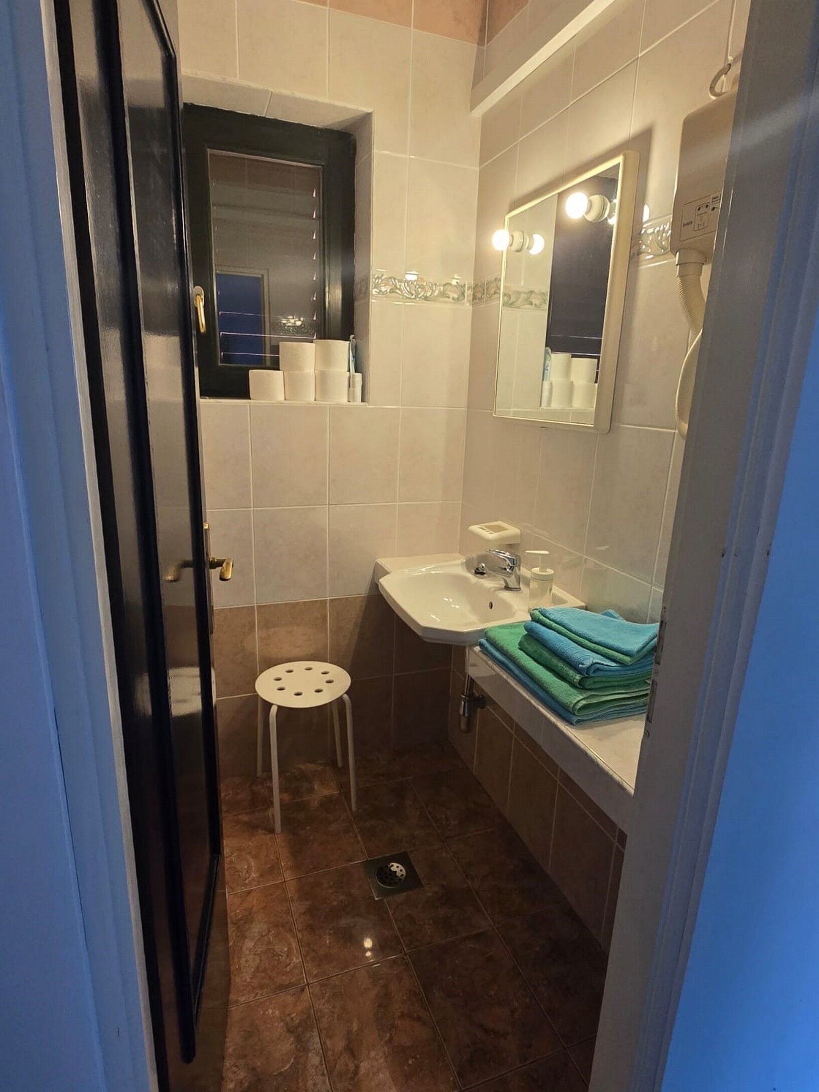 Ein kleines Badezimmer mit Waschbecken, Spiegel und Toilette. Handtücher liegen in der Nähe des Waschbeckens gestapelt und ein kleiner Hocker steht neben der Toilette.