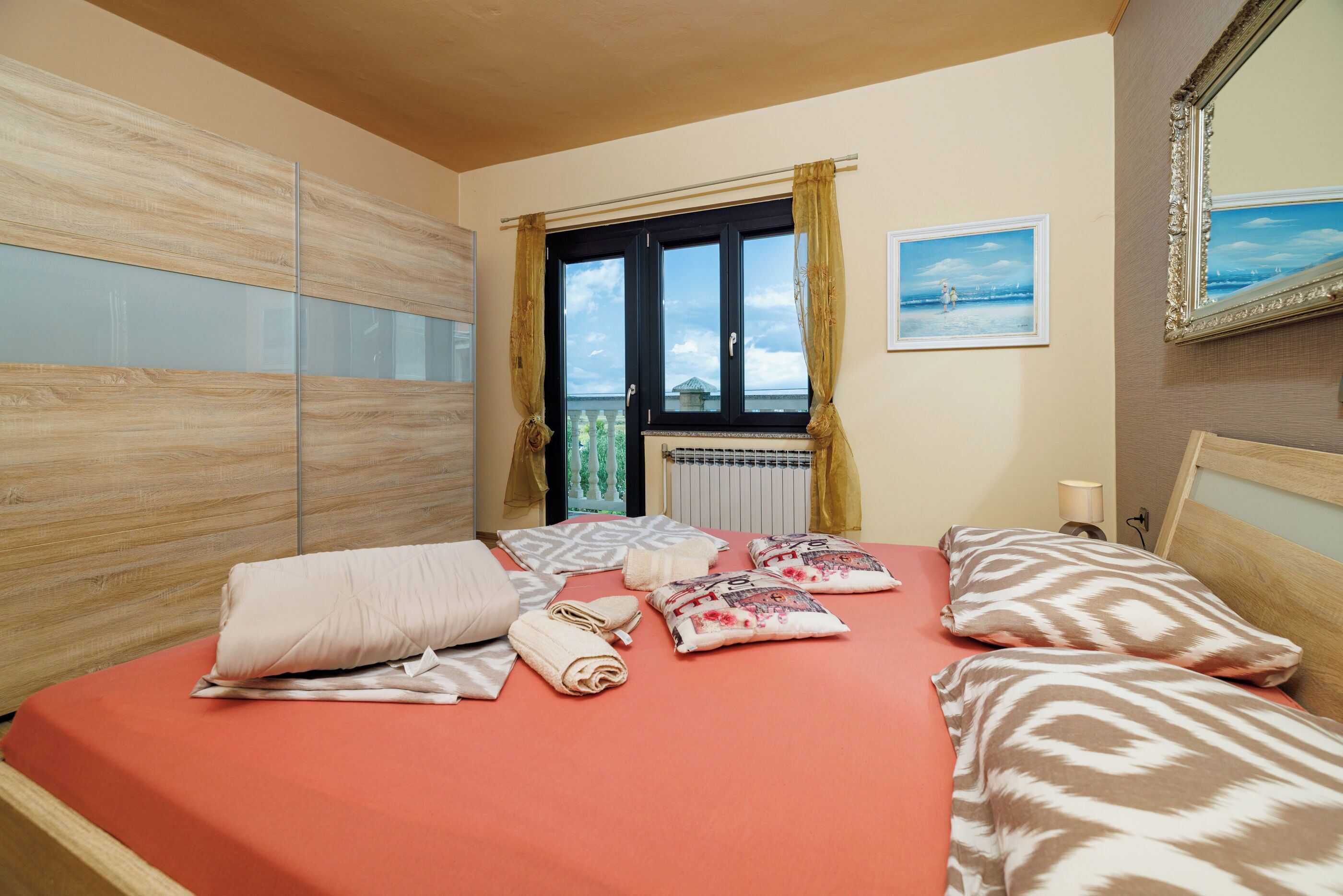 Une chambre d’hôtel avec un lit, des oreillers, une fenêtre avec des rideaux, un radiateur, un tableau accroché au mur et un miroir.