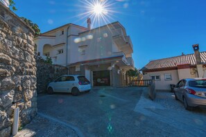 A street with two cars parked on the side, a stone wall on the left, and a building with a red-tiled roof in the background. - Apartment Vidas (Rab)