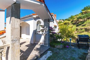 A white building with a stone foundation, a covered outdoor area with a barbecue grill, and a hillside with vegetation. - Apartment Vidas (Rab)