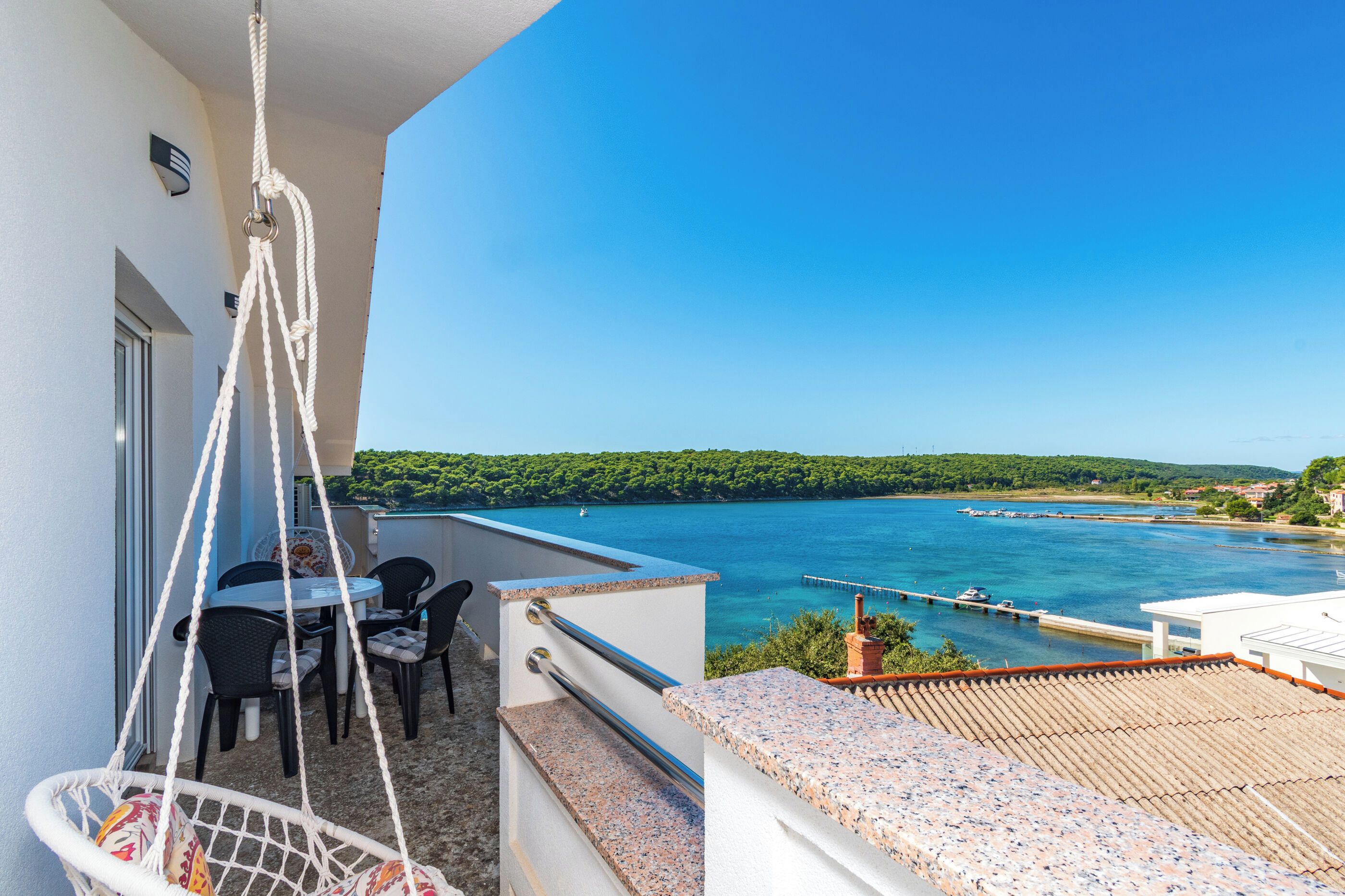 A balcony with a table and chairs, a hanging chair, and a view of a lake with boats and a pier.