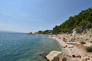 A beach with clear water, people sunbathing, and a rocky hillside with trees. - Apartment Lele (Omis)