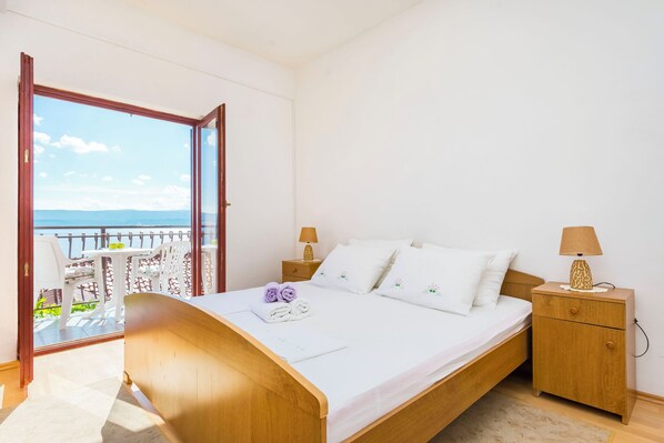 A bedroom with a wooden bed, white bedding, two bedside tables with lamps, and a balcony with outdoor furniture. - Apartment Lele (Omis)