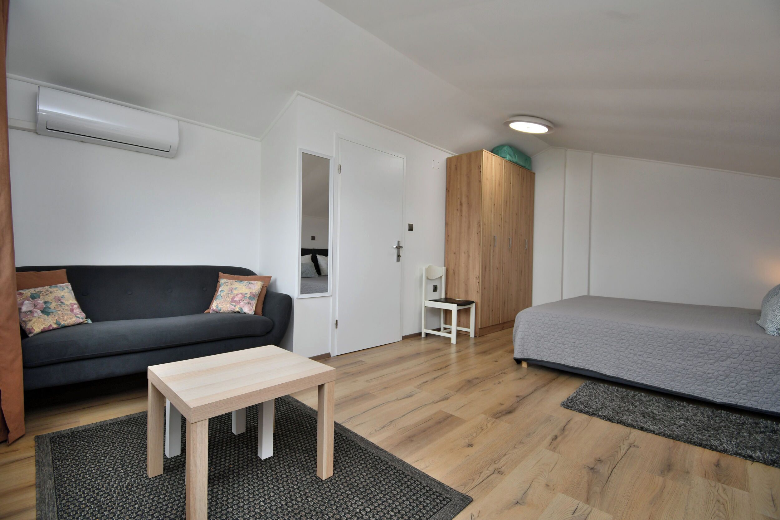 A modern, compact room with a grey sofa, a wooden table, a grey bed, and a wooden wardrobe.