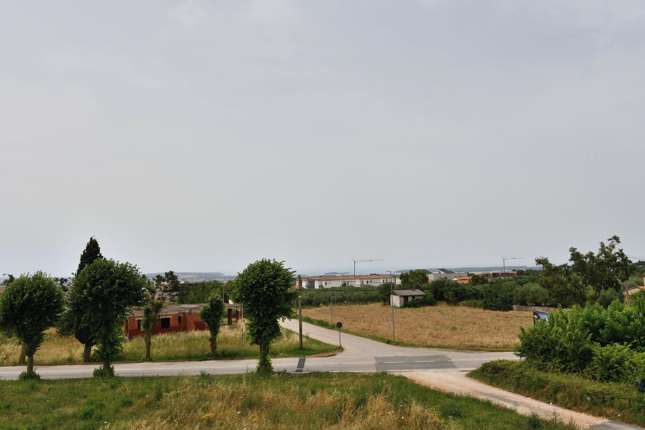 Een landweg met een bocht, omringd door groen en enkele gebouwen in de verte.