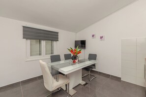 A modern dining area with a glass table, grey chairs, a vase with flowers, a flat-screen TV, and a window with blinds. - Apartments Beachfront Srima (Vodice)