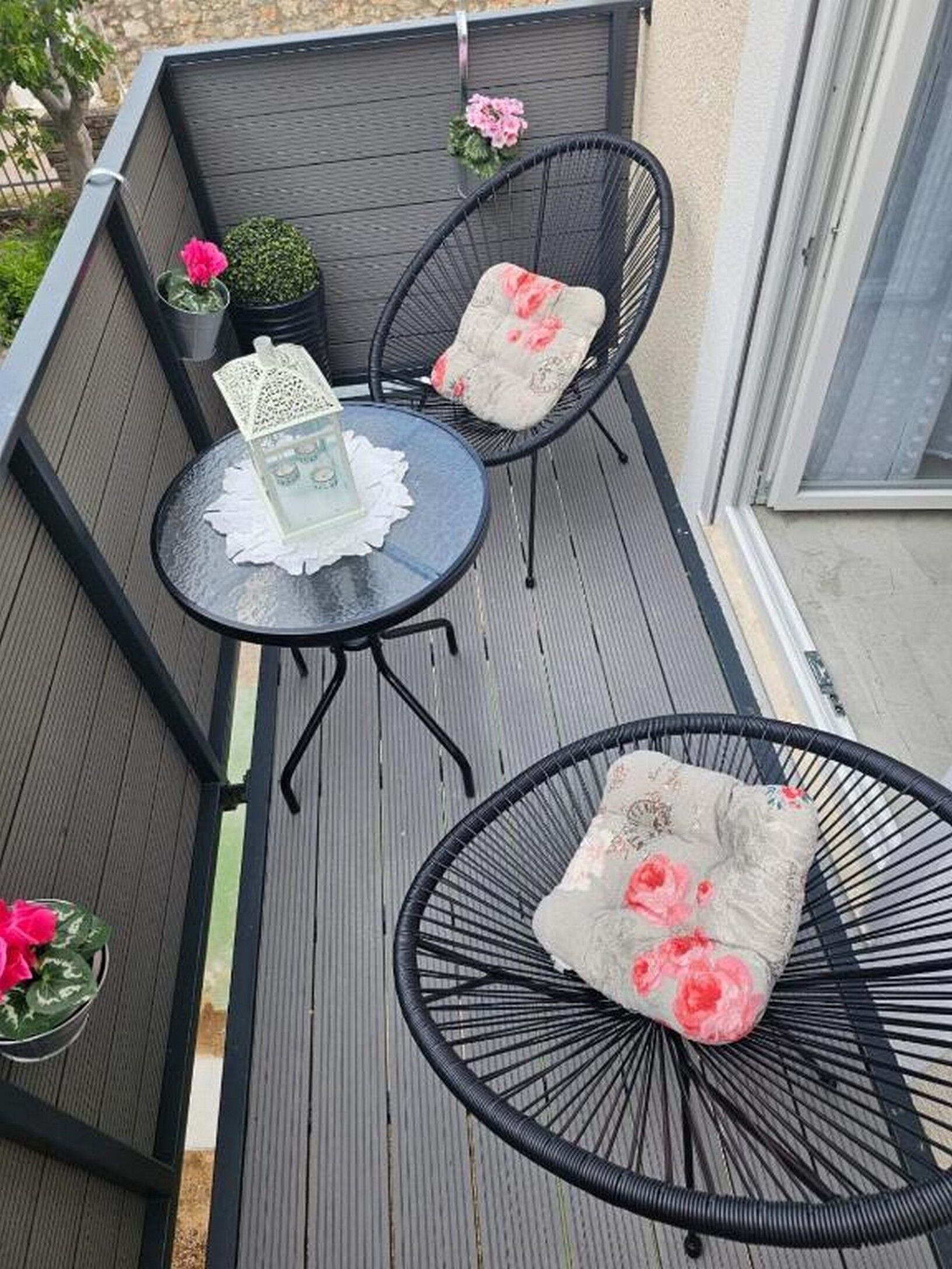 A small outdoor balcony with two black wicker chairs, a round table with a white lace doily, and potted plants with pink flowers.