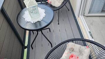 A small outdoor balcony with two black wicker chairs, a round table with a white lace doily, and potted plants with pink flowers.